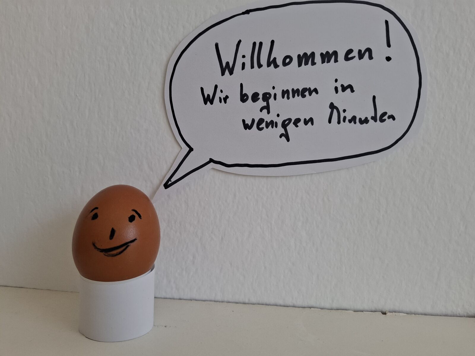 Ei mit aufgemaltem Gesicht im Eierbecher, daneben eine Sprechblase: „Willkommen! Wir beginnen in wenigen Minuten“